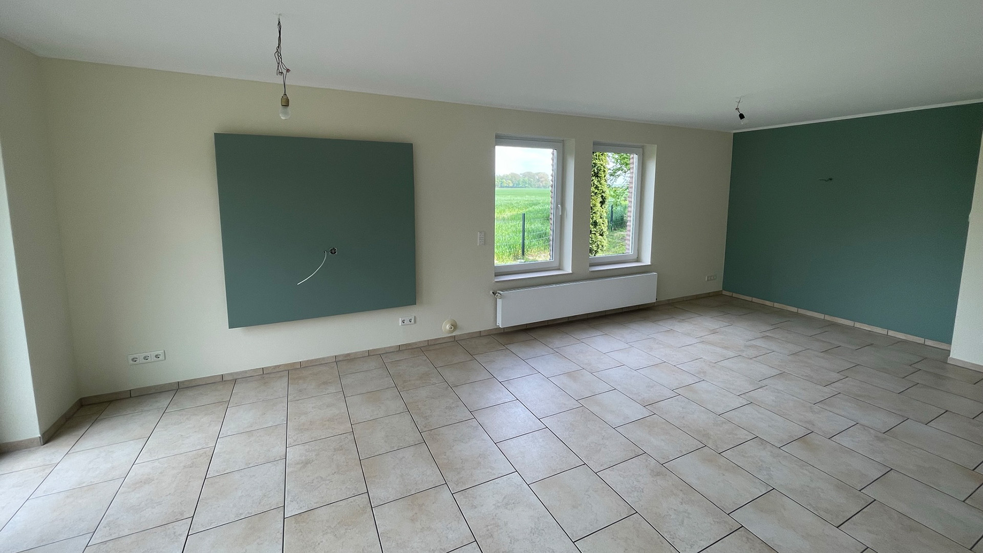 Leerer Raum mit beigen Fliesen, grüner Wandfarbe, zwei Fenstern und unvollendeter Beleuchtung.