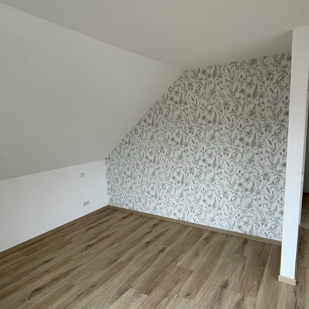 Leerer Raum mit gemusterten Tapeten und Holzfußboden, keine Personen sichtbar.