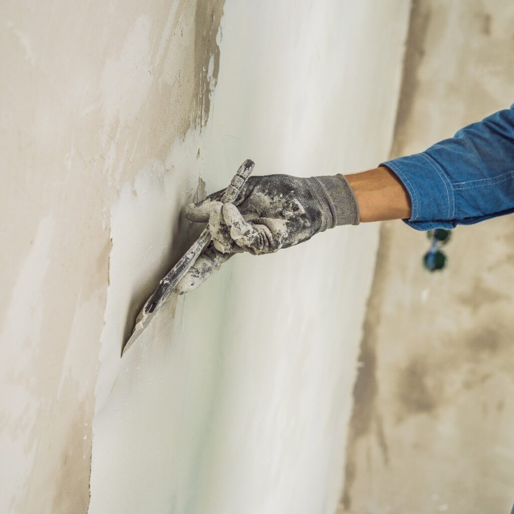 Meister trägt weiße Spachtelmasse auf eine Wand auf und verstreicht sie mit einem Spachtel in einem Raum eines renovierten Hauses bei Tag